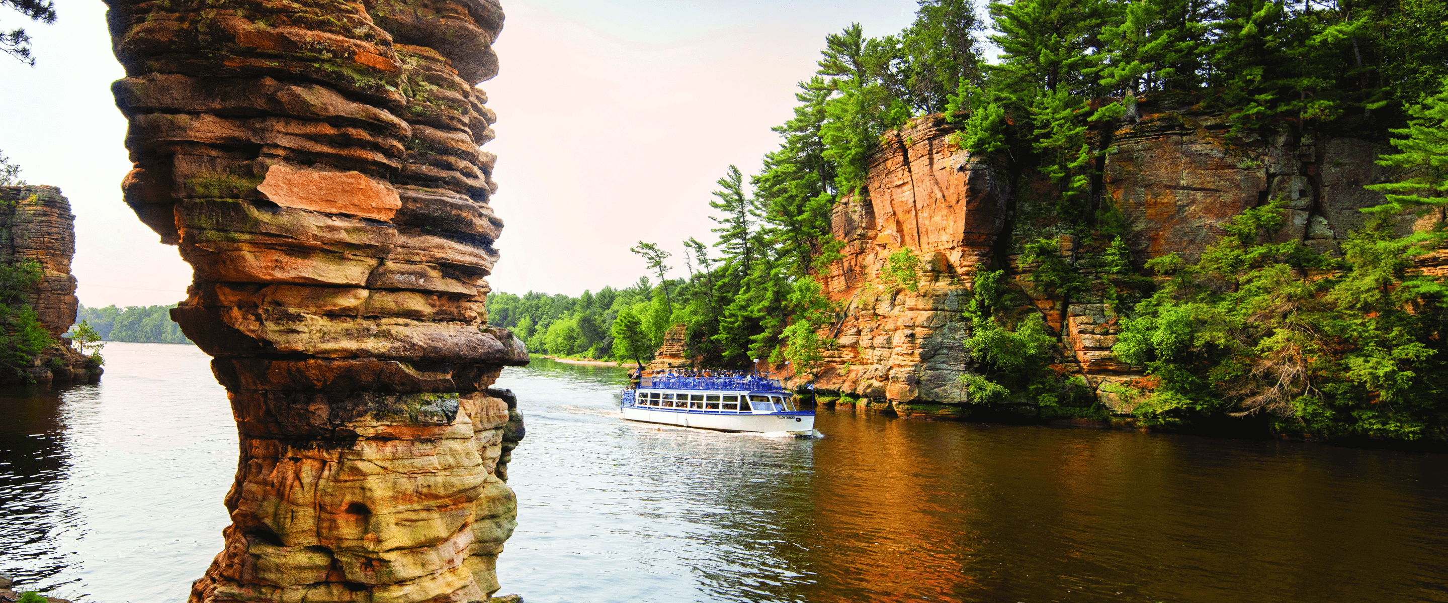 A blue tour boat rides among tall layered rocks and greenery in a waterway.